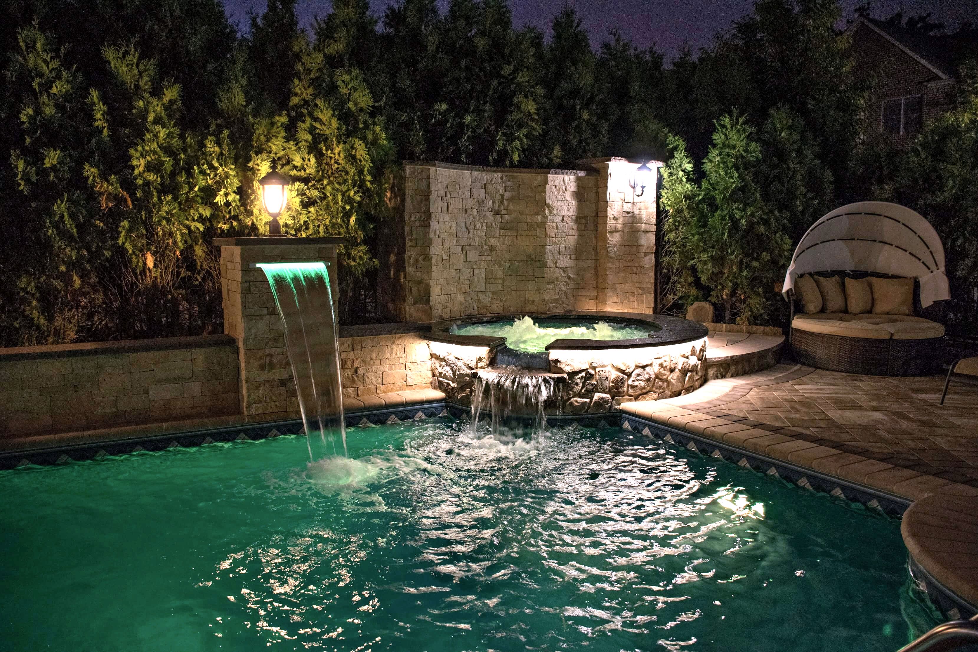 Night view of pool with green-lit waterfall, spa, and daybed lounge